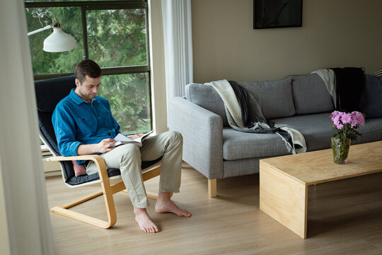 Man in 30s sitting on chair in living room reading document holding pen, copy space