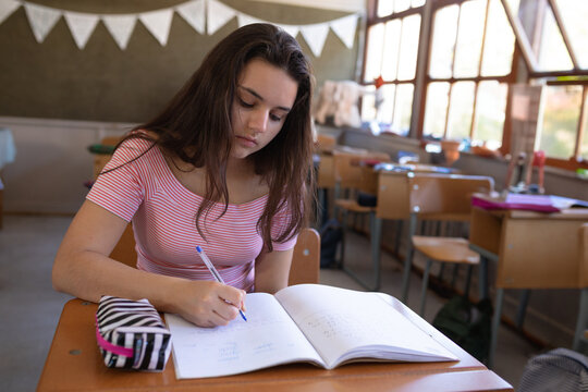 Teenage girl writing in notebook at wooden desk in classroom with blue pen, striped pencil case