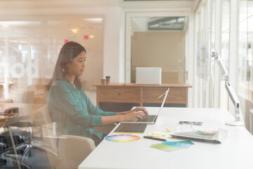 Asian woman typing on open laptop at modern office desk with color wheel, smartphone, copy space