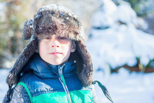 Boy standing amid snowy clearing with evergreen trees wearing puffy coat and trapper hat