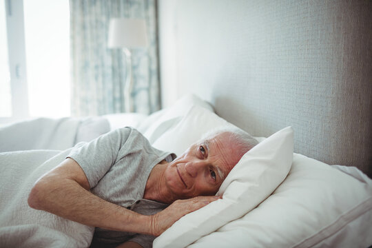 Senior man reclining on bed with white duvet and pillow beside window with patterned curtains