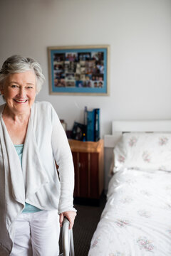 Senior woman leaning on walker in bedroom next to bed with floral bedspread, copy space
