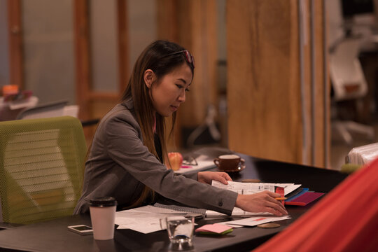 Asian woman wearing grey blazer sorting documents and color swatches at office desk with smartphone