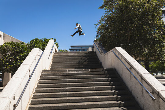 Man in sportswear leaping over gap on concrete stairs, railings, office block, trees in city plaza