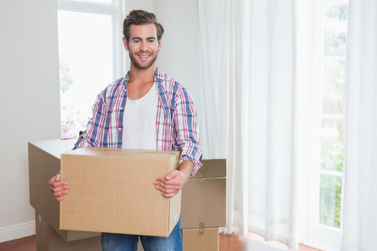 Man holding cardboard box unpacking in bright living room with stacked boxes and sheer curtains