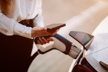 Woman charging electric car in parking lot during sunset with mobile device in hand
