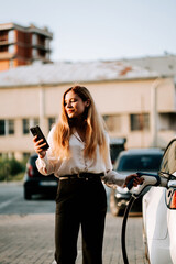 Woman charges electric car in parking lot at sunset while interacting with her smartphone