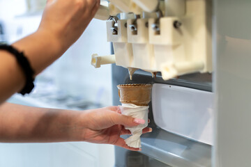 Delicious Treat Preparation: Close-up of female hand holding crispy waffle cone receiving a perfectly formed creamy swirl of mixed chocolate and vanilla soft serve ice cream from dispenser machine.