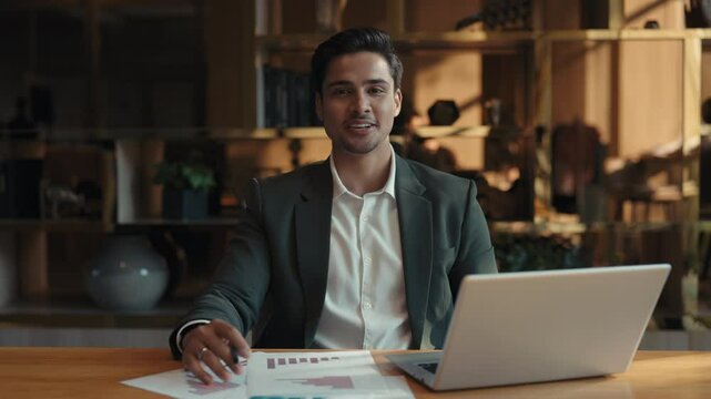 Portrait of positive Indian business consultant with laptop presents course in office camera view. Young businessman records tutorial for entrepreneurs sitting in co-working room