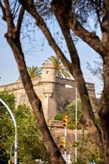 Fototapeta premium A historic stone fortress with a cylindrical turret rises behind trees and traffic lights, framed by lush palm fronds and warm daylight; fortress; architecture.