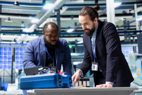 Multiethnic business associates and engineers gather for a discussion in a solar tech manufacturing facility, planning future developments in sustainable energy and industrial operations.