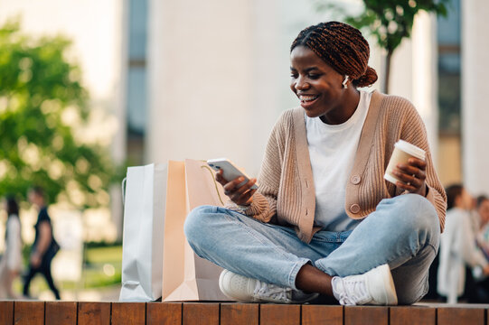 Smiling shopper using smartphone and holding coffee with shopping bags