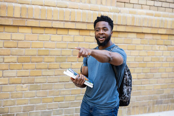 A cheerful young man pointing enthusiastically at something while holding books, exuding...