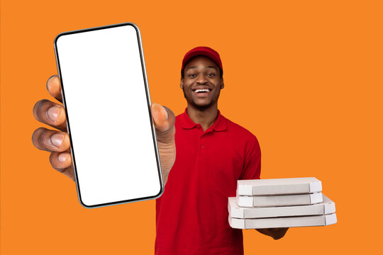 Fototapeta Fast Service And Best Food Delivery During Pandemic. Positive black courier wearing red uniform and cap holding pizza boxes from restaurant and showing okay sign gesture, isolated on orange background
