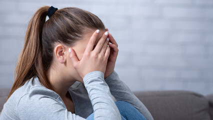 Side view of young woman with depression covering her face and crying on sofa at home. Upset...