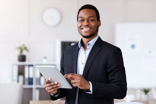 Smiling black entrepreneur young man holding digital tablet, using modern technologies in business, office interior, copy space. Cheerful african american businessman using app for business