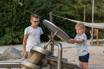 Focused young boys playing with interactive water screw pump at outdoor playground on sunny day. Concept of childhood curiosity, teamwork, educational play, and public park activities.