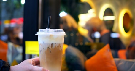 Barista presenting a delicious iced coffee with whipped cream and a straw, in a transparent plastic cup, with blurred customers and orange pillows in the background