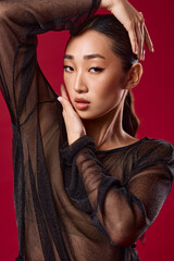 Asian woman in sheer top posing with hands on head against red background, hair pulled back