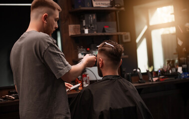 Barber cuts a man's hair with clippers in a cozy, sunlit barbershop. The client sits with clips in...