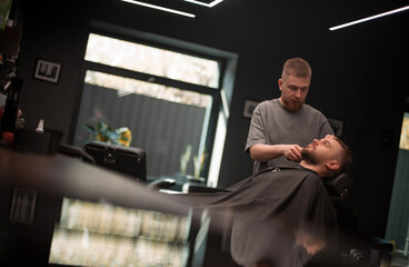 A young barber gently trims the beard of a relaxed male client reclining in a modern barbershop....