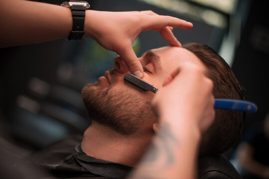 Close-up of a professional barber using a straight razor to shape a client's beard while the man relaxes with closed eyes during a grooming session in a modern barbershop..