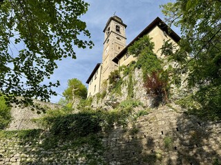 View from below of the Sanctuary of Madonna del Soccorso in Ossuccio, Lake Como, Italy, with stone walls, bell tower and lush greenery under a bright blue spring sky