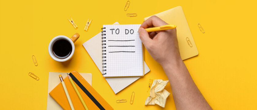 Male hand with to-do list, notebooks, cup of coffee and stationery on yellow background