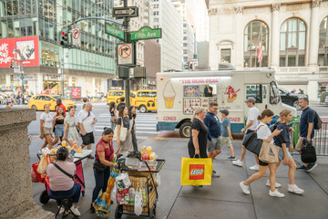 Obraz premium New York City, New York, USA - 07.29.2024: People walk past an ice cream truck on the corner of Fifth Avenue and West 42nd Street in New York City. Street vendors and shoppers with colorful bags add