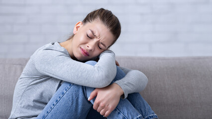 Mental health care concept. Young depressed woman sitting on sofa with closed eyes and hugging her...