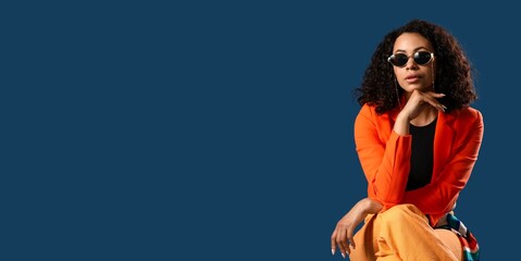Stylish young African-American woman in orange blazer and with sunglasses on dark blue background...