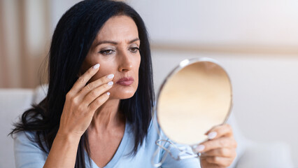 Aged Skin. Portrait of serious senior woman with long dark black hair holding mirror and looking at...