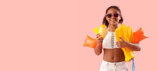 African-American girl with bottles of sunscreen cream, swimming armbands and flippers on pink background with space for text © Pixel-Shot