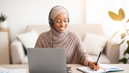 Black Muslim lady in hijab and headset having video conference on laptop, taking notes during...