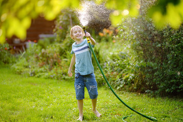 Funny preteen boy pouring cool water on himself and playing with garden hose with sprinkler in sunny backyard on hot summer day. Happy child having fun with water splashes.