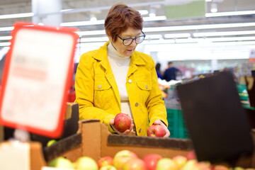 Beautiful red-haired senior lady is choosing fresh organic apples in a food store or a supermarket .