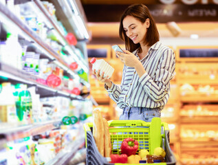 Smiling young woman taking dairy products from shelf in the supermarket, holding bottle and smartphone, scanning bar code on product through mobile phone, walking with trolley cart