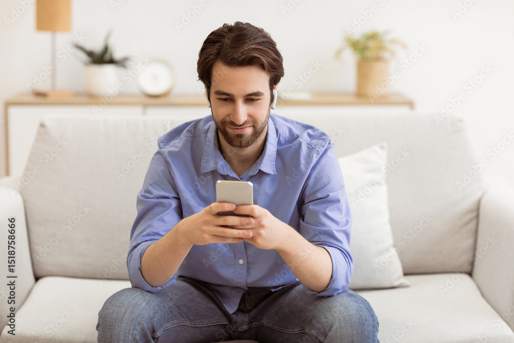 Poster man with earbuds using phone browsing internet and listening to music online sitting on couch at hom