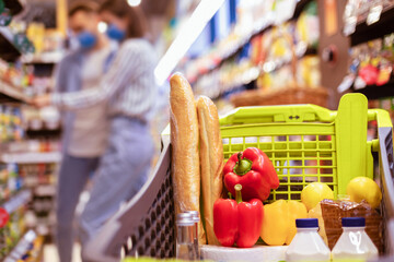 Family Shopping And Healthy Products. Young Couple Standing In Supermarket In The Blurred Background, Selective Focus On Shopping Trolley Cart Full Of Food, Vegetables, Bread And Dairy