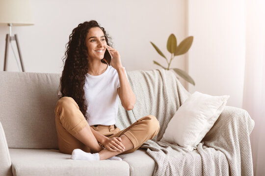 Happy brunette woman with long curly hair having conversation on mobile phone while sitting on couch in living room and looking at copy space. Young lady talking on phone and smiling