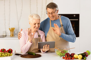 Happy Senior Couple Video Calling Using Digital Tablet, Waving Hand To Computer Webcam Standing In Modern Kitchen Cooking Together At Home. Virtual Remote Communication By Video Call Concept