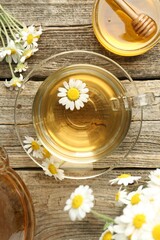Delicious chamomile tea in glass cup, fresh flowers and honey on wooden table, flat lay