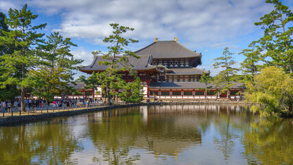 Nara, Japan - Sep 26 2024, Panoramic view across the pond and trees to the facade of Todaiji Temple, Great Buddha Hall, with a blue cloudy sky, with people on background, Nara, Japan