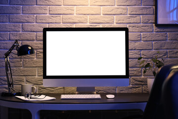 Blank computer monitor with glowing lamp on desk in dark office at night