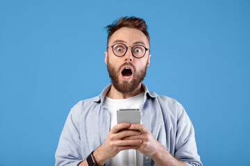 Shocked bearded guy staring at mobile phone in disbelief over blue studio background. Surprised young man with smartphone learning unbelievable news, amazed at huge online sale