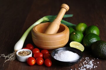 Fresh ingredients including green onions, cherry tomatoes, limes, an avocado, coarse salt, mixed spices, and a wooden mortar and pestle on a dark wooden surface