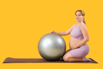 Sporty young pregnant woman with fitball sitting on yoga mat against yellow background