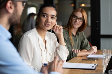 Three young professionals having a discussion around a wooden table in a modern office setting, showing engagement and thoughtful expressions