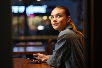 Young woman with ponytail sitting at a wooden table in a dimly lit cafe holding a smartphone and looking thoughtfully upward