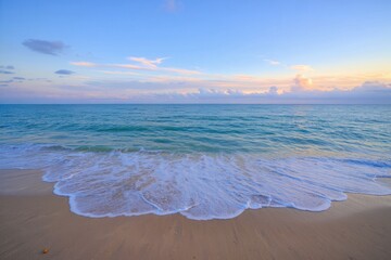 Gentle ocean waves rolling onto a sandy beach under a clear sky with soft clouds during sunset, creating a peaceful and serene atmosphere
